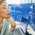 A serious woman with short curly hair, wearing clear glasses and a light-colored top, sits at a desk with a laptop and multiple computer screens displaying financial data and graphs