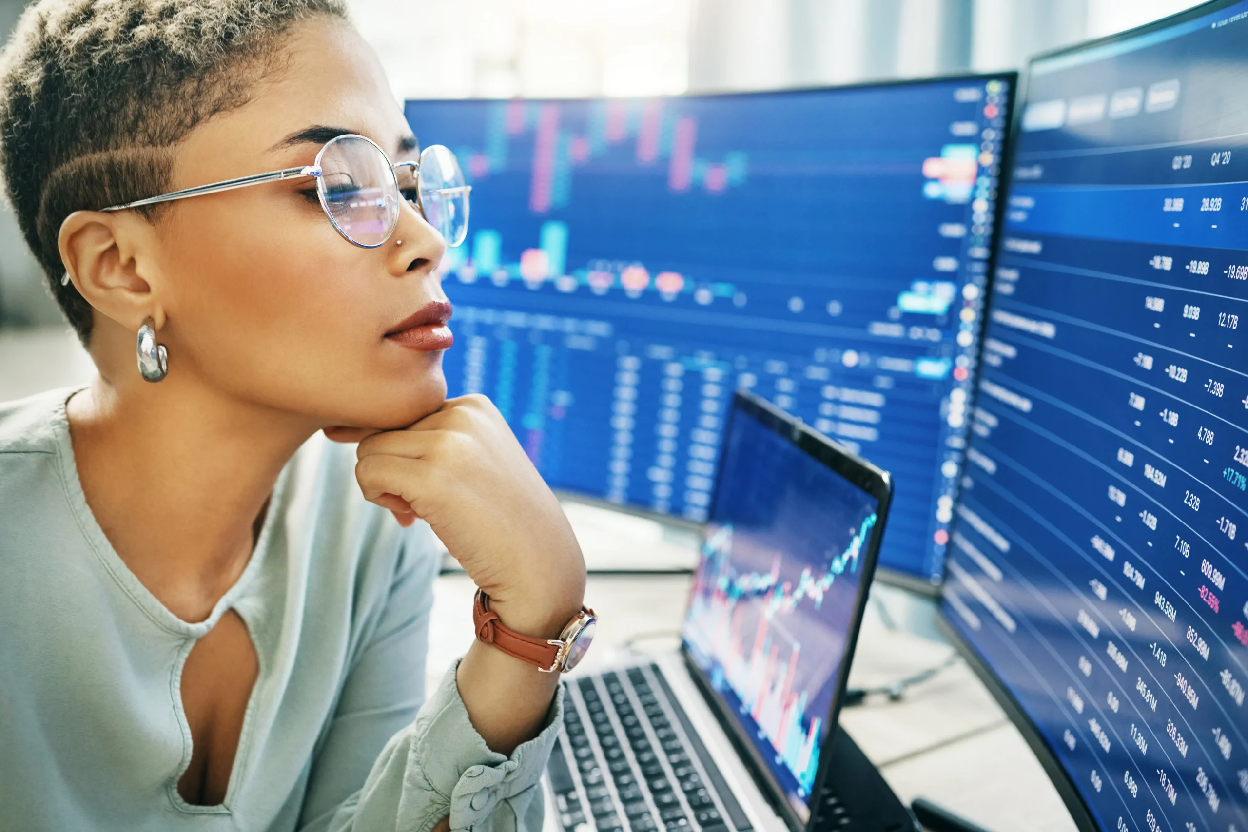 A serious woman with short curly hair, wearing clear glasses and a light-colored top, sits at a desk with a laptop and multiple computer screens displaying financial data and graphs