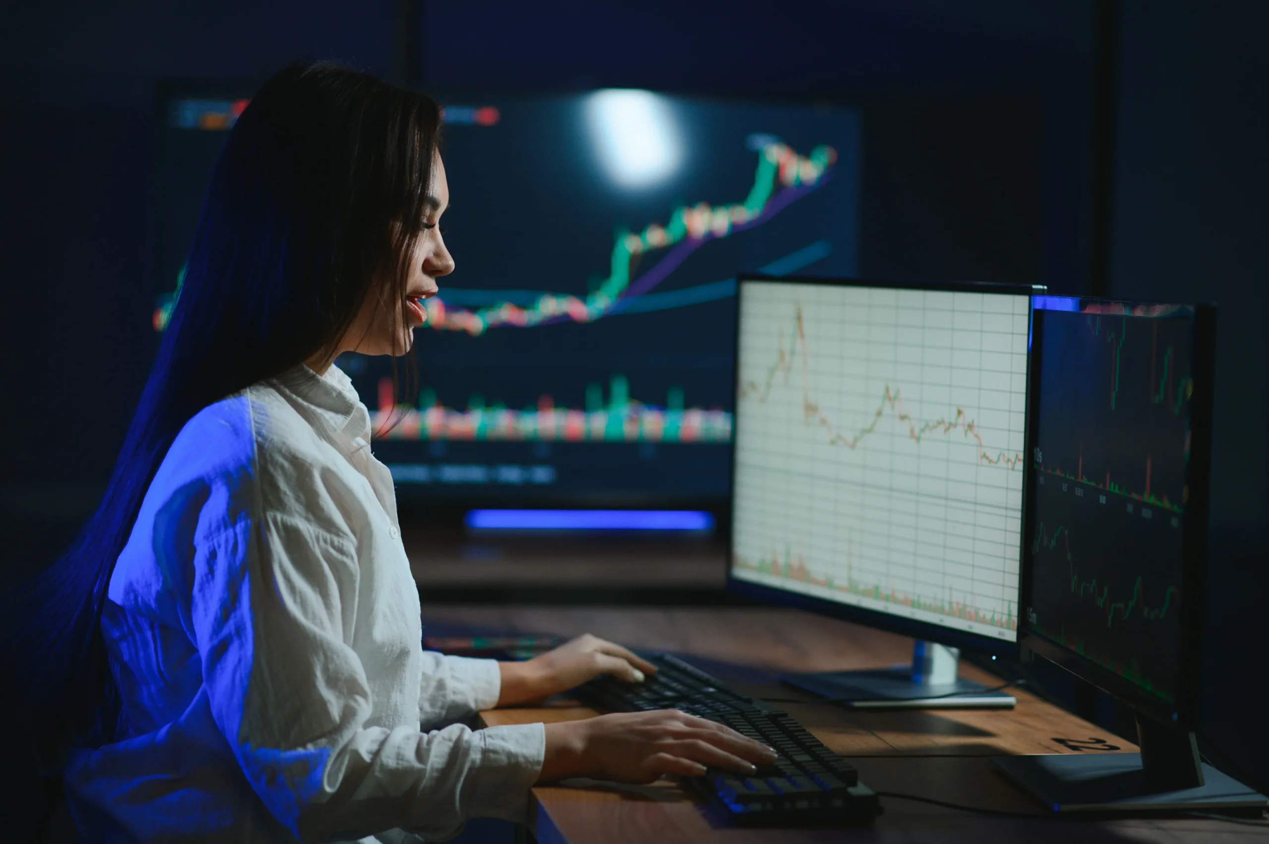 A woman with long dark hair is seated at a desk, intently focused on multiple computer screens displaying charts and graphs about the stock market