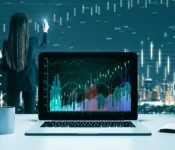 An adult with long dark hair analyzes data on a laptop. The woman is seen from the back, and wears a business suit. She holds a pen to the transparent display, seemingly annotating the data shown.