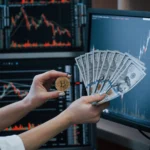 Close-up view of hands holding a gold-colored coin with the bitcoin logo next to a fanned stack of United States one hundred dollar bills. The background shows computer monitors displaying stock market charts with fluctuating graphs in red and white on a dark background. The image suggests concepts related to finance, investment, and cryptocurrency. Ideal for projects centered on business, investment, economics, and modern financial technologies.