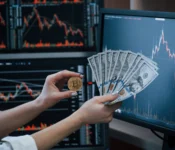 Close-up view of hands holding a gold-colored coin with the bitcoin logo next to a fanned stack of United States one hundred dollar bills. The background shows computer monitors displaying stock market charts with fluctuating graphs in red and white on a dark background. The image suggests concepts related to finance, investment, and cryptocurrency. Ideal for projects centered on business, investment, economics, and modern financial technologies.