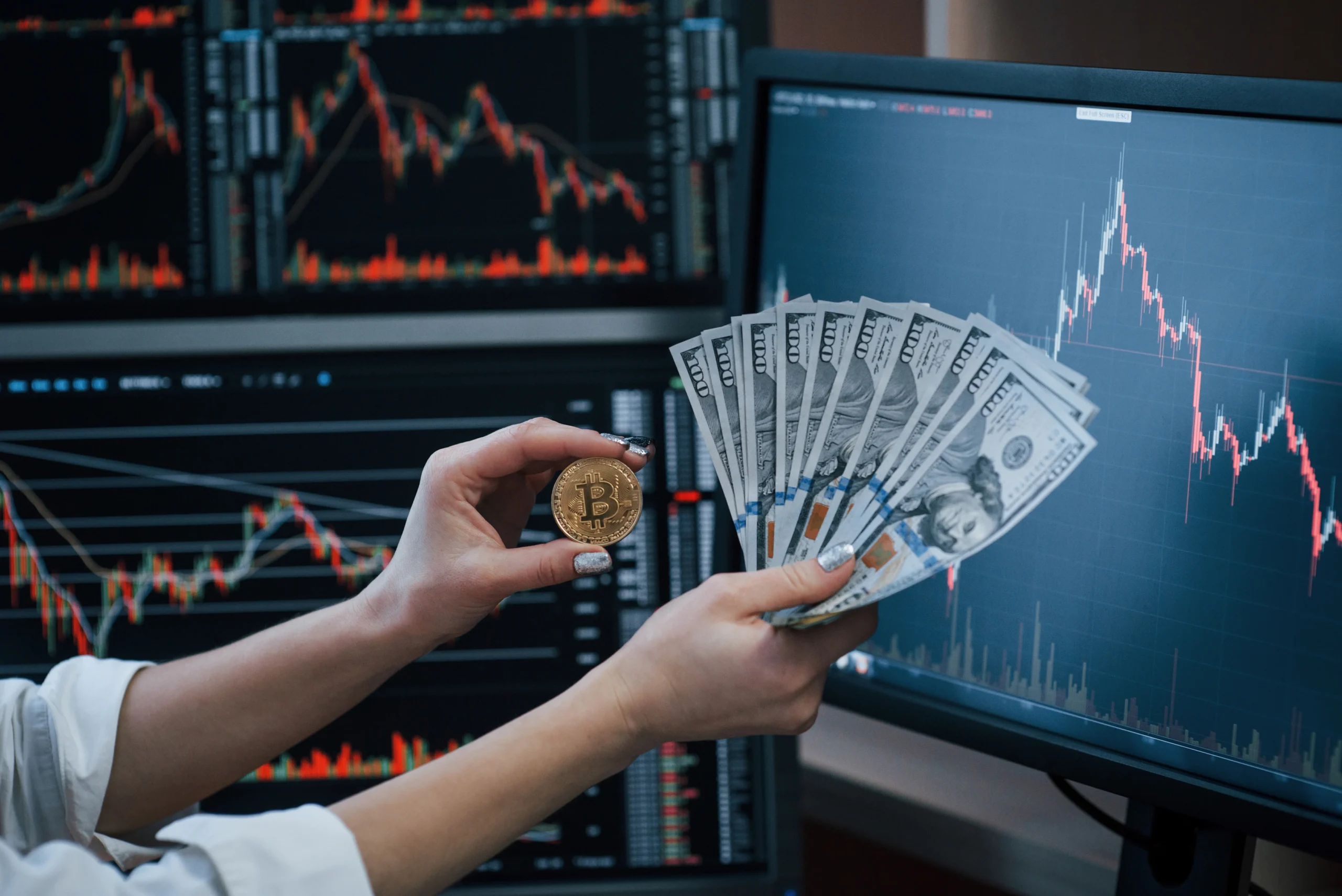 Close-up view of hands holding a gold-colored coin with the bitcoin logo next to a fanned stack of United States one hundred dollar bills. The background shows computer monitors displaying stock market charts with fluctuating graphs in red and white on a dark background. The image suggests concepts related to finance, investment, and cryptocurrency. Ideal for projects centered on business, investment, economics, and modern financial technologies.