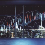 Imagery shows financial stock market charts overlaid onto a large window. Through the window a city skyline is visible at night. Graph lines illustrate the rise and fall of data. The blue and orange graphs on the window and the blurred city lights in the background.