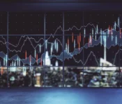 Imagery shows financial stock market charts overlaid onto a large window. Through the window a city skyline is visible at night. Graph lines illustrate the rise and fall of data. The blue and orange graphs on the window and the blurred city lights in the background.