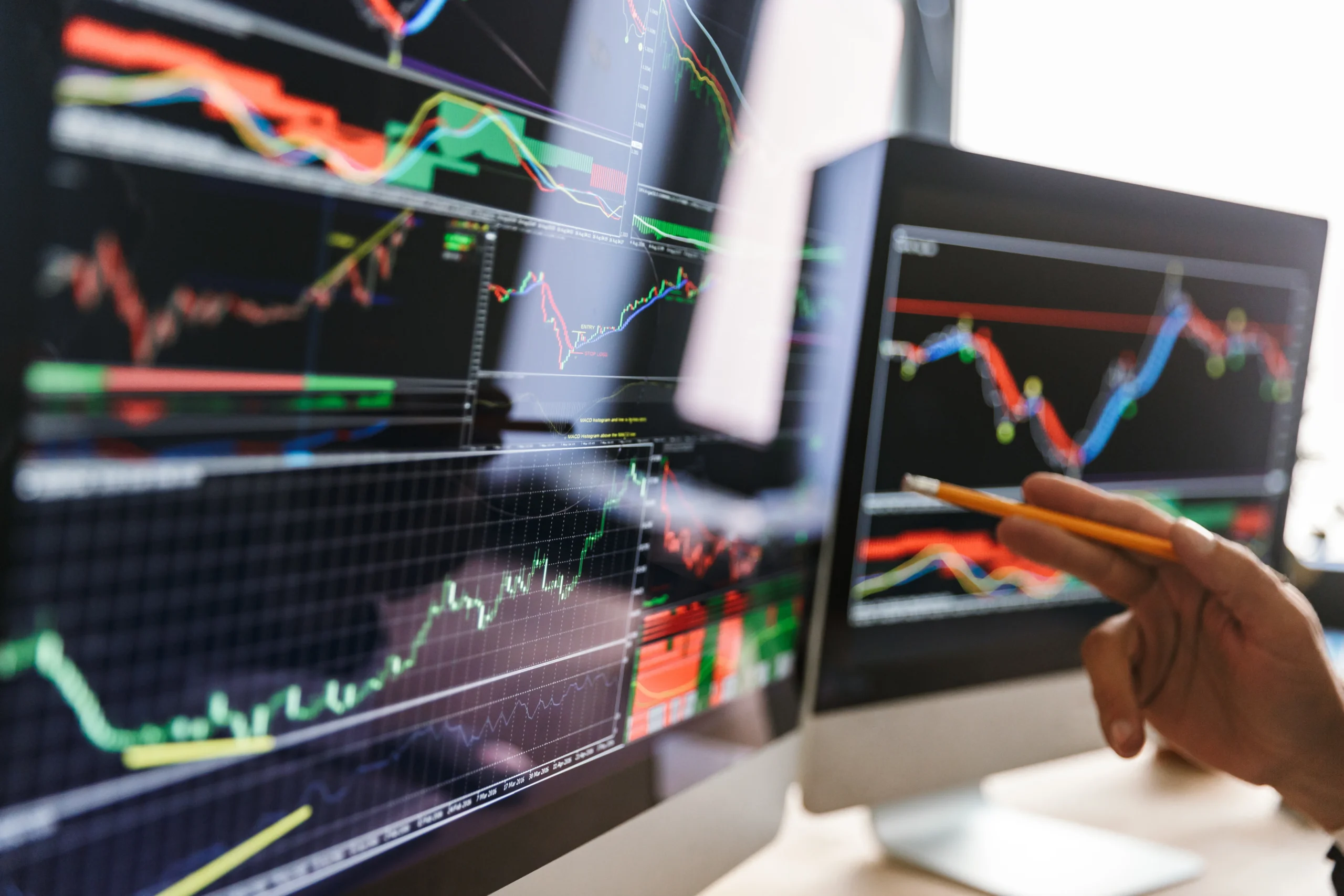 The image shows a close-up of two computer monitors displaying stock data with upward trending lines, indicating progress and gain. An arm and hand are partially visible, with the hand holding a pencil and appearing to analyze the data on screen. The setting appears to be an urban workplace, with bright daytime lighting. This compelling photo could be used for projects related to technology, investment, business, finance, and corporate success.