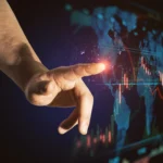 A disembodied hand is seen touching a glowing spot on a digital world map, with financial graphs displayed. The map features blue lines and red financial charting that suggests a futuristic, technology-driven environment. The background is a dark blue with a high contrast with shadows.