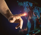 A disembodied hand is seen touching a glowing spot on a digital world map, with financial graphs displayed. The map features blue lines and red financial charting that suggests a futuristic, technology-driven environment. The background is a dark blue with a high contrast with shadows.