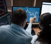 Two financial analysts, a man and a woman, are reviewing data displayed on multiple computer monitors. The monitors show charts and graphs with fluctuating lines.