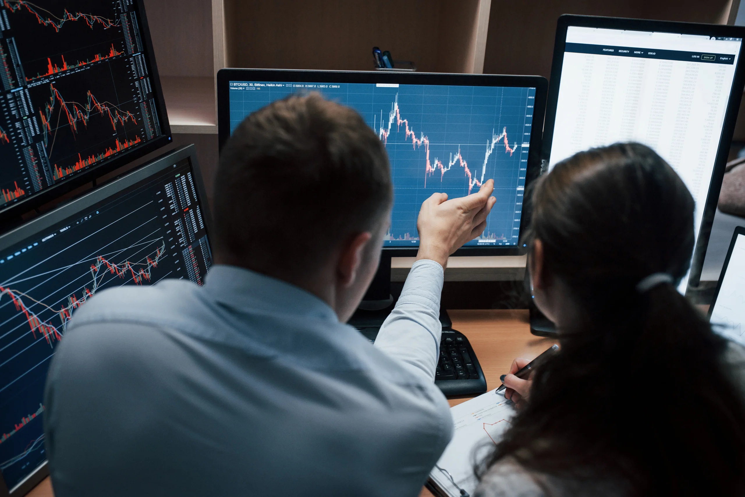 Two financial analysts, a man and a woman, are reviewing data displayed on multiple computer monitors. The monitors show charts and graphs with fluctuating lines.